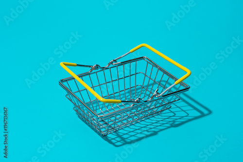 Empty metal shopping basket with yellow handles on bright turquoise blue background. Concepts of retail, shopping, consumer choice and sale season.