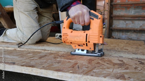 A jigsaw in the hand of a worker kneeling on a thick sheet of OSB and cutting the dense material into pieces, the sharp blade of the jigsaw cuts the massive OSB sheet