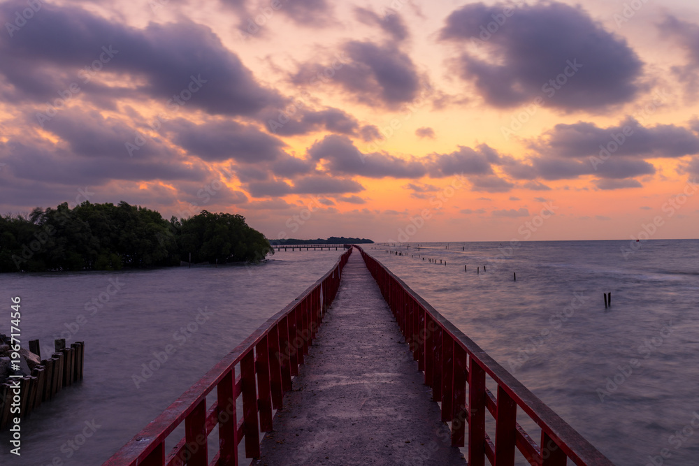 Obraz premium A view of the red bridge crossing the sea at sunrise,A stunning long-exposure photograph featuring a vibrant red wooden bridge stretching toward the horizon over the calm sea. The scene is captured.