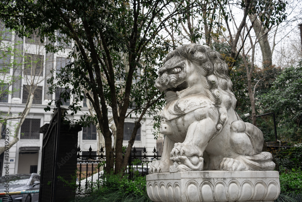 Obraz premium Stone lion sculpture in a courtyard with green trees and architectural background