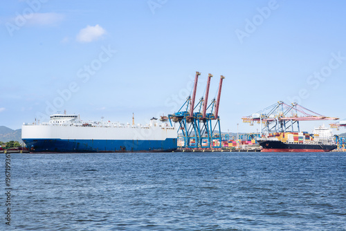 Car carrier ship docked at port terminal in Brazil being loaded with vehicles, while tugboat passes nearby, maritime logistics concept.