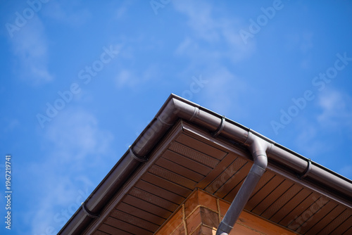 Brown gutter system on roof corner of wooden house against blue sky. Exterior drainage installation for rainwater management. Concept of home maintenance and residential construction.