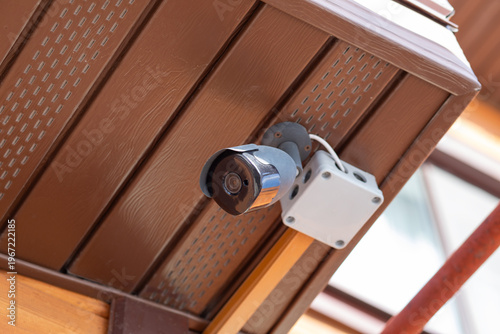 Security camera mounted on brown ceiling under roof. Surveillance system for property protection and safety monitoring. Digital video equipment for prevention of burglary and crime control.