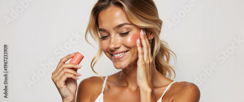 Young woman applying cream blush stick on her cheek while smiling  