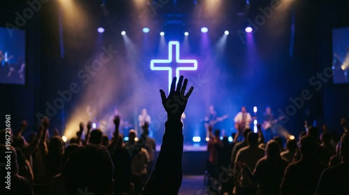 Silhouetted hand raised in worship before a glowing neon cross. Christian concert prayer service at church. Spiritual praise and adoration during Easter Sunday worship celebration. 