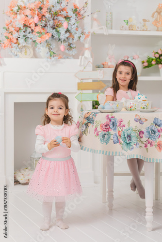 little girl holding easter egg in bright spring interior with another child at festive table, soft pastel decor and blooming flowers creating joyful holiday mood