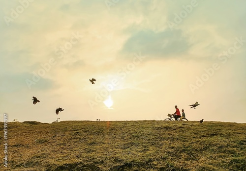 Silhouette of Two People Riding Bicycle on Grassy Hill with Birds Flying at Sunset Golden Hour Lighting Pastoral Scene