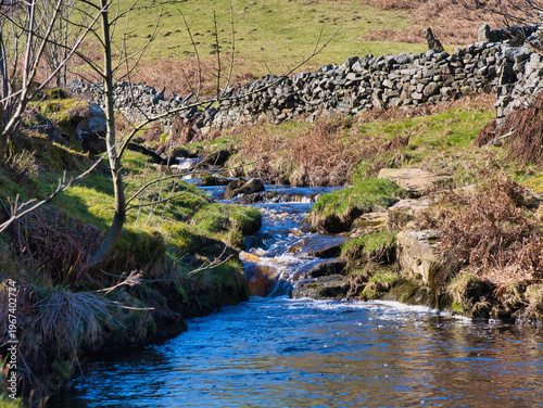 A clear rural stream winds through winter grass and rocks beside a dry stone wall in Yorkshire UK, with bright sunlight and a crisp countryside feel