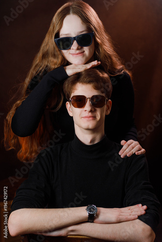 Cool siblings or friends in dark outfits with sunglasses against brown background.