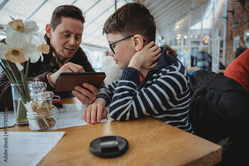 Father showing tablet to son sitting at table indoors learning together