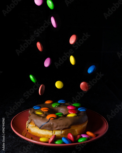 Chocolate glazed donut with colorful candy falling on dark background