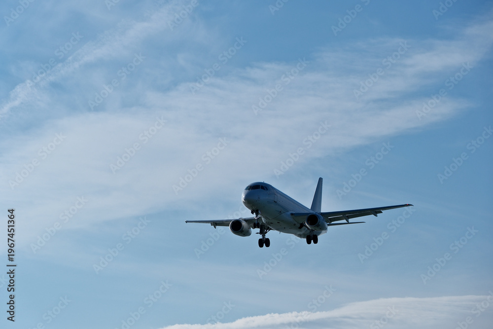 Obraz premium White passenger plane on final approach on a sunny day with blue sky, light clouds and beautiful backlighting. Sunlit white airplane landing against a blue sky with wispy clouds, backlit by bright sun