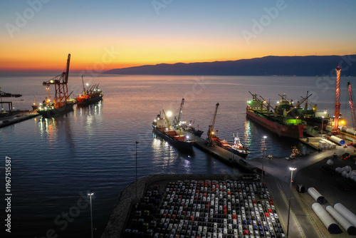 A bustling commercial port where illuminated cargo ships dock, cranes operate under a colorful sunset sky, and a vast parking lot is filled with new cars awaiting export.