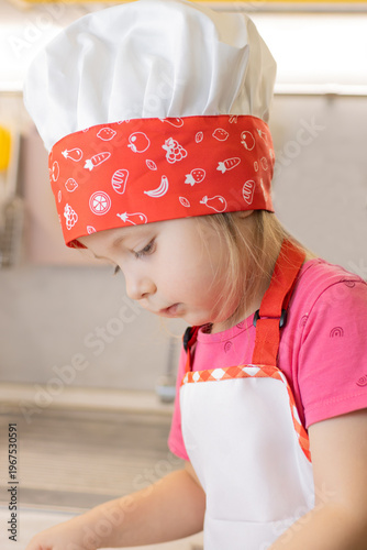 Little girl chef in a colorful fruit-patterned hat prepares to cook in the kitchen. This photo captures joyful children's cooking moments perfectly.