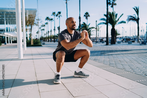 Fotografía Smiling man doing deep squats in tropical city setting with palms, lower body strength training, joy of outdoor fitness, healthy habits, authentic representation, copy space, looking away