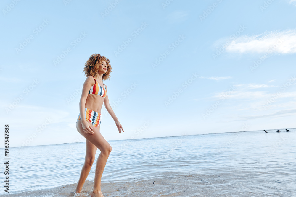 Naklejka premium Young woman walking on beach shore in colorful swimsuit, enjoying sunny day near calm sea, relaxed lifestyle, natural light, clear sky, summer vacation concept