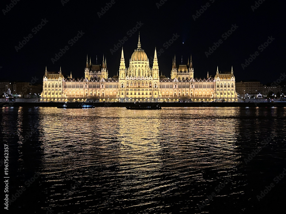 Fototapeta premium Night view of the Hungarian Parliament building in Budapest with reflections on the Danube River and illuminated historic architecture.