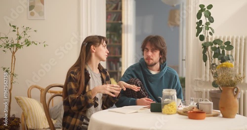 Young couple sitting at kitchen table during breakfast, discussing plans and writing in notebook in cozy sunlit apartment