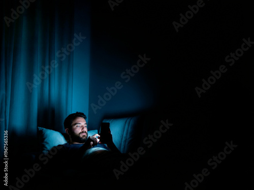 Man uses smartphone in dark room while lying on bed at night, focused on screen and surrounded by shadows
