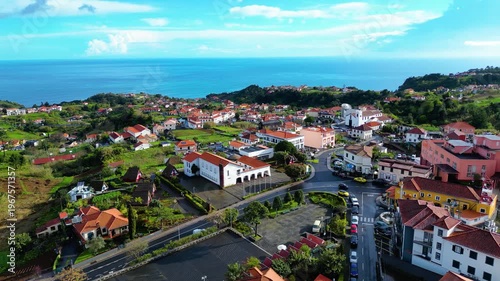 Aerial view of Santana, Madeira, showcasing its iconic red-roofed houses, lush hillsides, and the vast Atlantic Ocean under a clear blue sky.