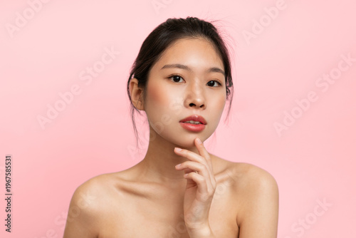 A young woman stands with a thoughtful look on her face. She has one finger on her chin and is facing the camera. The background is a solid pink color, creating a bright atmosphere.