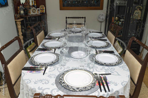 Elegant dining table set for eight with white tablecloth, silver chargers, glassware and cutlery arranged for formal meal at home