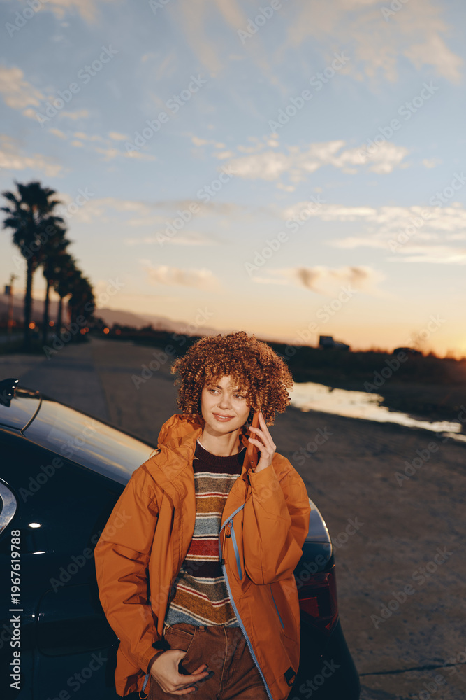 Fototapeta premium Young woman smiling outdoors near car during sunset wearing rainbow sweater and orange jacket, holding smartphone, relaxed lifestyle, casual autumn outfit, natural light