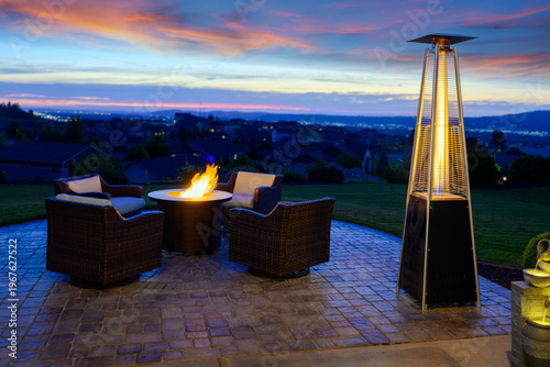 Fotografie A Pyramid patio heater and flaming fire pit warm a residential paver tile terrace with seating on a cold evening sunset, overlooking a suburban neighborhood and city lights