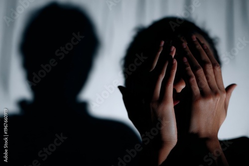 A person covers their face with both hands as a shadow looms on the wall