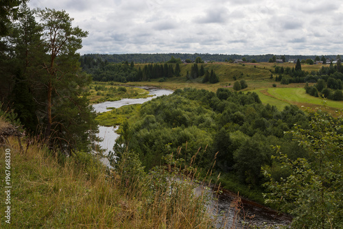 A view from a high bank of a river bend and a village. Summer rural landscape. In the distance, on the opposite bank of the Gorodishna River, there are wooden houses. Norovo, Vologda Region, Russia.