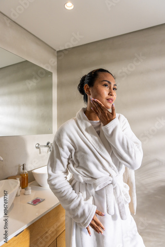 Asian woman wearing bathrobe, standing in bathroom beside vanity and mirror, checking skincare