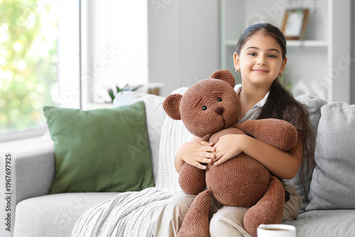 Little girl with toy bear sitting on sofa at home