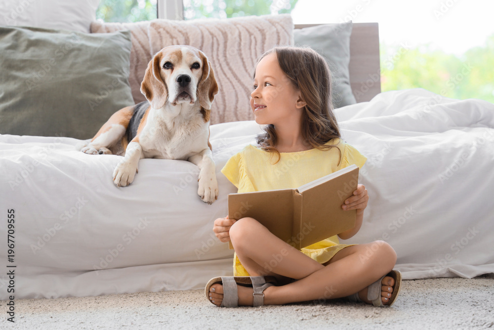 Obraz premium Cute little girl with Beagle dog reading book near bed in bedroom