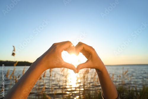 Bild auf Leinwand Female hands making heart gesture against river at sunset