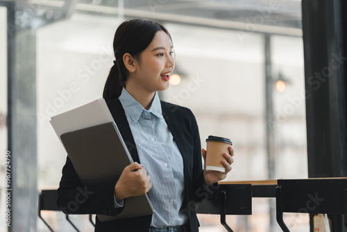 Foto Asian business woman holding laptop and coffee cup standing in modern office