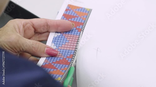 Horizontal close-up shot of an artisan's hand holding a printed technical pattern for bead weaving.