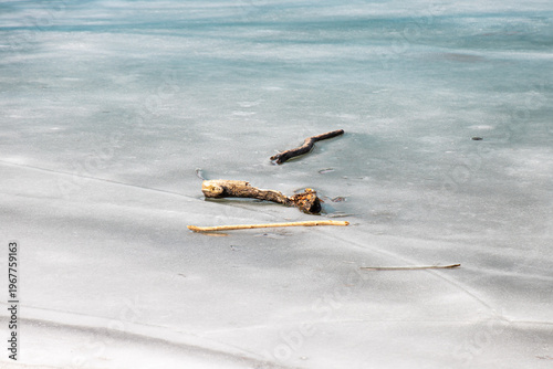 Driftwood branches trapped in the pale blue-grey ice of a frozen lake, with open water visible in the background.