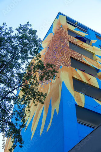 Colorful mural facade on modern apartment building, abstract sunflower pattern, blue yellow shapes rising against sky expressing urban art creativity architecture identity, positive city atmosphere