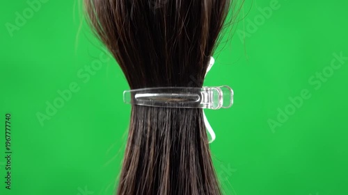 A clear plastic hair clip secures a section of dark brown hair against a vibrant green background, highlighting a simple accessory for hair styling