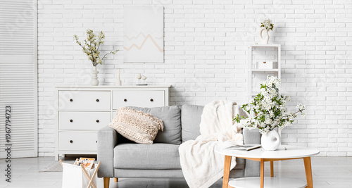 Sofa with dresser, coffee table and vase of blooming branches near white brick wall in living room