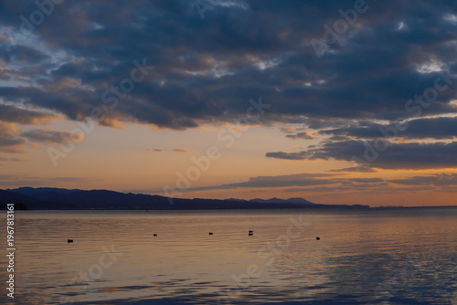 夕暮れの湖面に映る空と遠景の山々、水鳥が静かに漂う