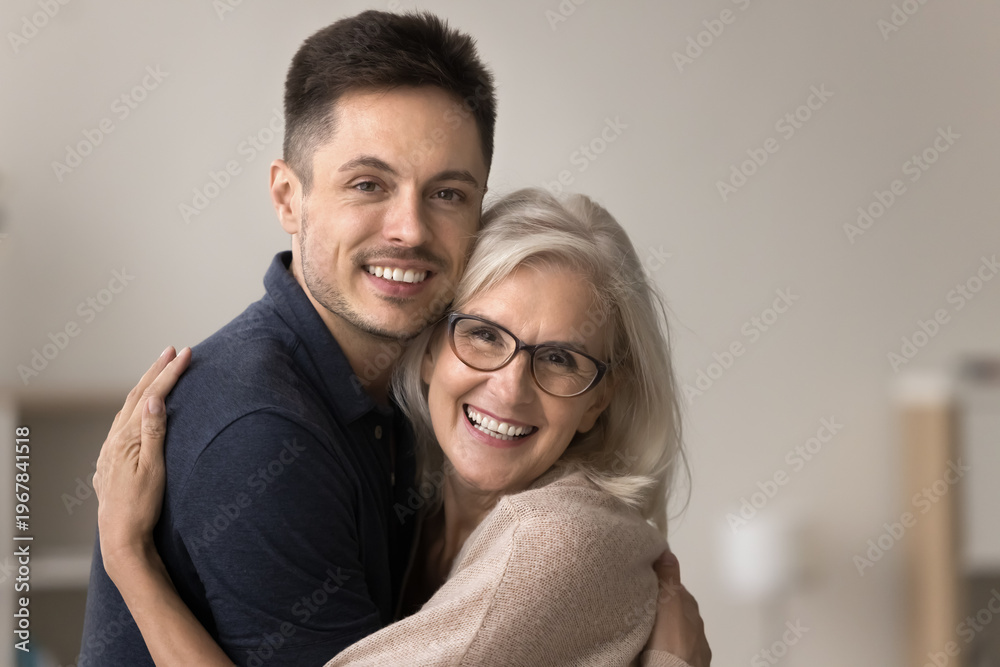 Obraz premium Close up faces, portrait of young adult man hugging tightly his smiling middle-aged mother showing love and care. Smiling grown-up son embrace senior mom, enjoy priceless family time at home together