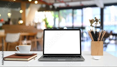 A laptop sits on a white table with a cup, notebooks, and pencils in a blurred cafe background with natural light.
