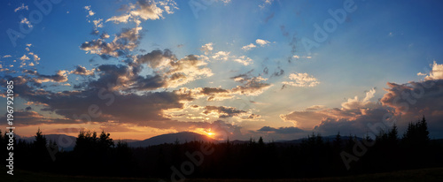 Serene evening sky with hues of blue and soft pink clouds as sun sets. Horizon glows gently, casting light over silhouetted pine trees and rolling hills, creating peaceful and expansive landscape.