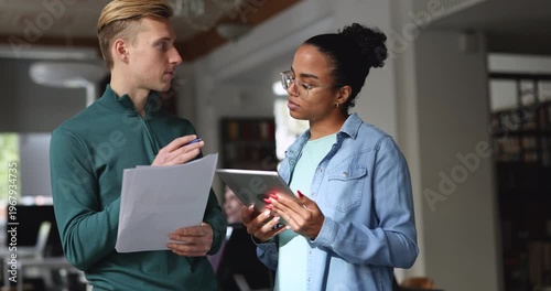 Two business colleagues standing in office reviewing documents, checking data on digital tablet, discussing report, working on project details, analyzing information. Collaborative workflow, teamwork