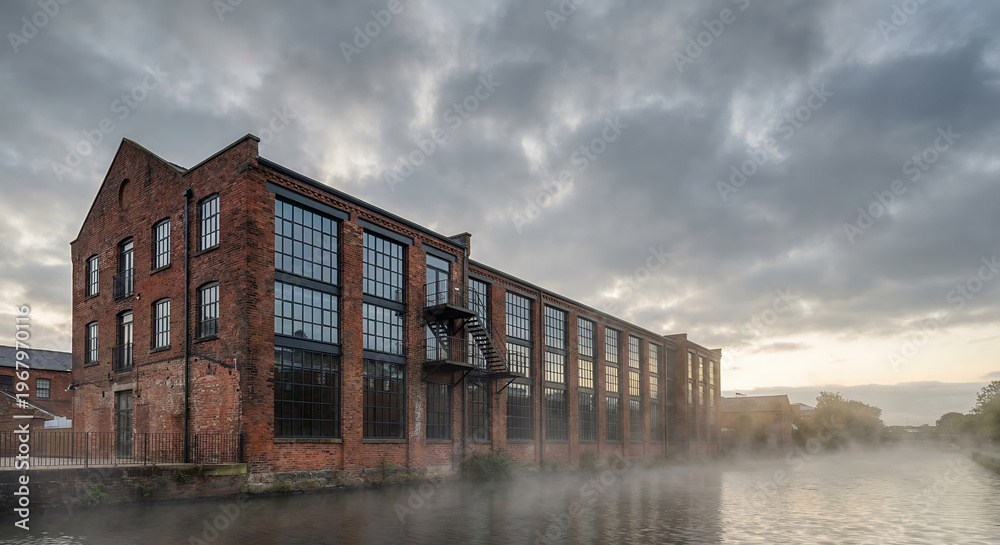 Fototapeta premium A brick industrial building with large windows stands beside a misty canal under a cloudy sky.