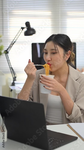 Vertical vdo woman eating instant noodles at desk, fast meal choice highlighting convenience versus long term health risks.