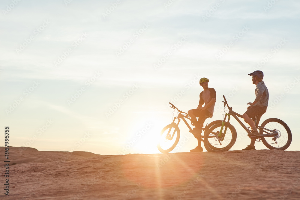 Fototapeta premium Men, mountain bike or rest with sunset in desert for extreme sport or fitness together in nature. Flare, male people or cyclist with friend or bicycle for cycling break or outdoor challenge on space