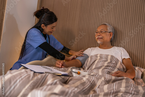 Caring young Asian female nurse checking blood pressure of happy elderly male patient resting peacefully in comfortable home bedroom during routine health medical checkup visiting patient