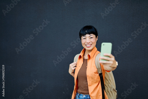Portrait of a happy asian woman posing, using a smartphone phone or taking a selfie photo outdoors in front of a modern urban background